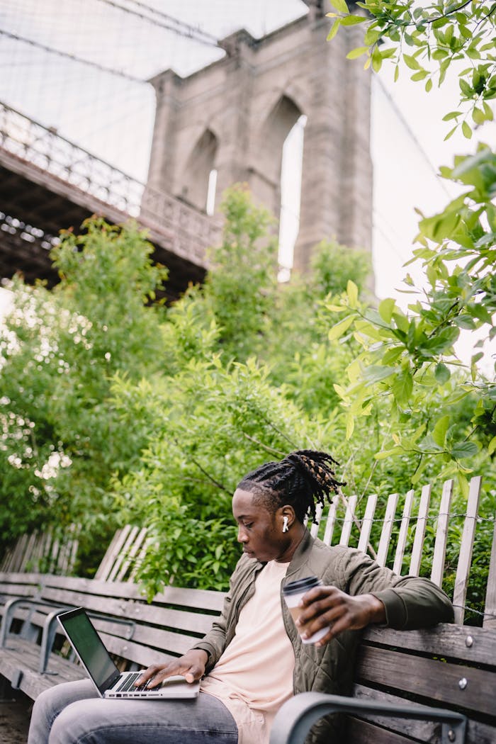 Side view of young African American guy with dreadlocks in casual outfit drinking coffee from disposable cup and typing on laptop while chilling on bench in city park